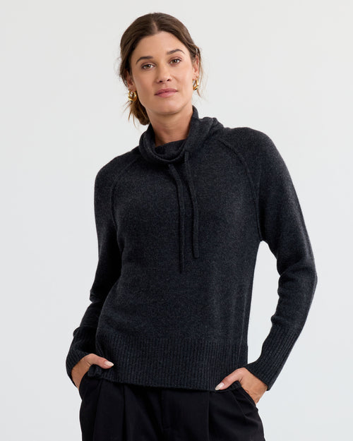Woman wearing charcoal cashmere sweater with cowl neck and drawstrings, hands in pockets, neutral background