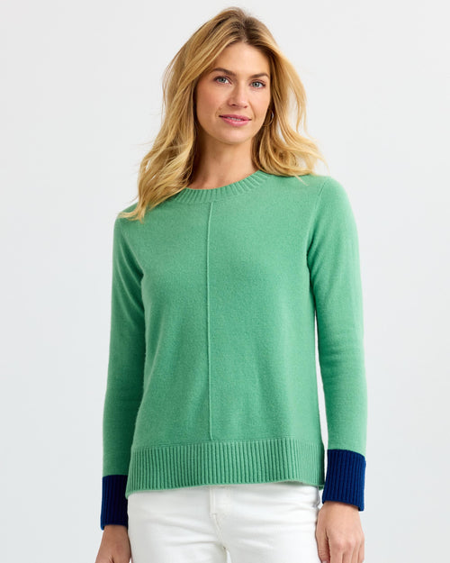 Woman wearing green cashmere sweater with navy cuffs and white pants against a plain light background