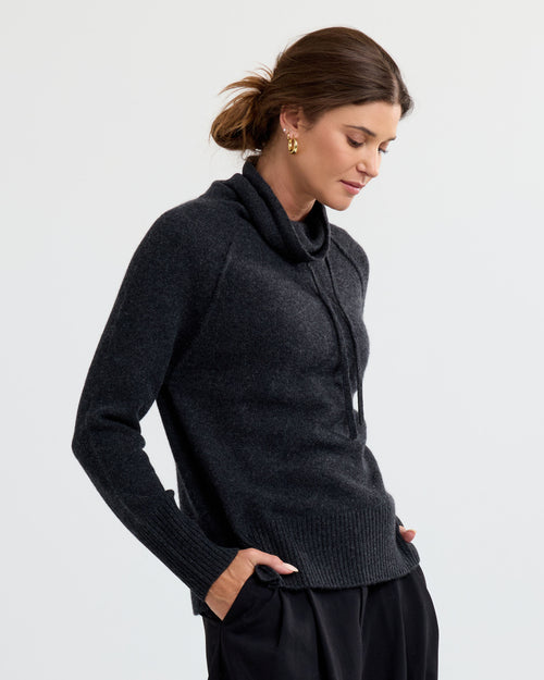 Woman wearing charcoal cashmere sweater with cowl neck and drawstrings, hands in pockets, against plain background