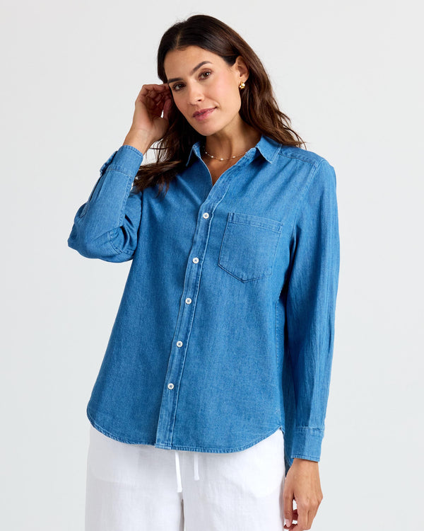 LIFESTYLE / EDITORIAL: Woman wearing medium wash denim shirt and white pants against plain background