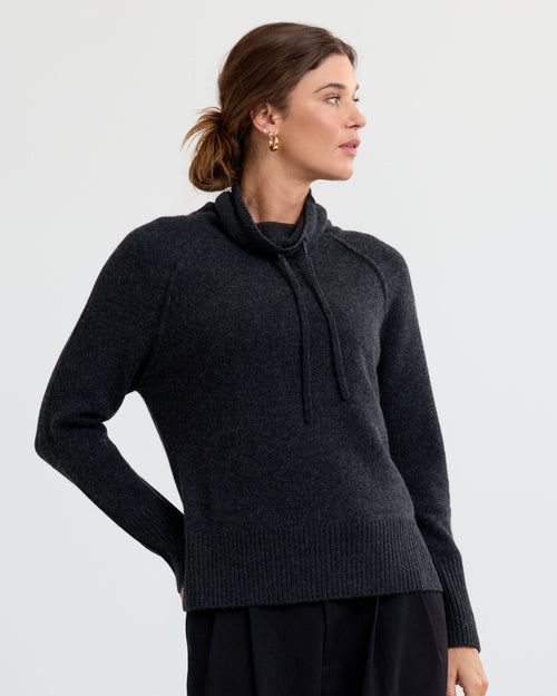 LIFESTYLE / EDITORIAL: Woman wearing charcoal cashmere sweater with drawstring collar, looking to side against plain