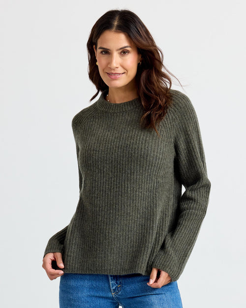 Front three-quarter image of a woman wearing a dark olive ribbed cashmere sweater with blue jeans