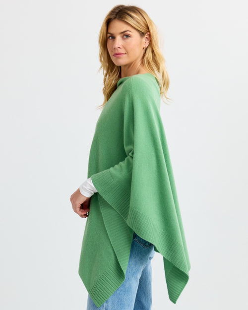 Woman wearing green cashmere poncho over white top and jeans, standing sideways against white background