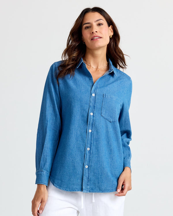 LIFESTYLE / EDITORIAL: Woman wearing medium wash blue denim shirt and white pants against a plain background