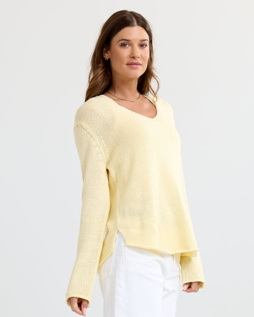 Woman wearing buttermilk yellow cashmere V-neck sweater and white pants, standing against plain background