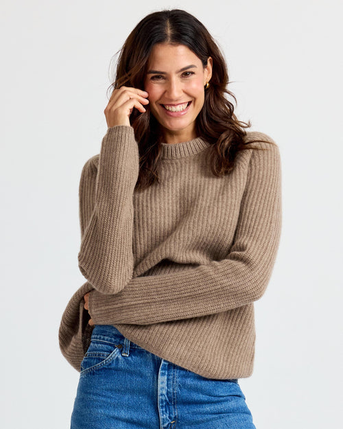 LIFESTYLE / EDITORIAL: Smiling woman wearing driftwood-colored ribbed cashmere sweater and blue jeans against white
