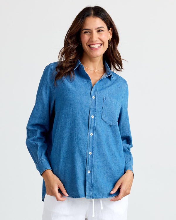 LIFESTYLE / EDITORIAL: Woman smiling wearing medium wash denim shirt and white pants against plain background