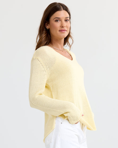 LIFESTYLE / EDITORIAL: Woman wearing a buttermilk yellow V-neck cashmere sweater and white pants, standing against a white