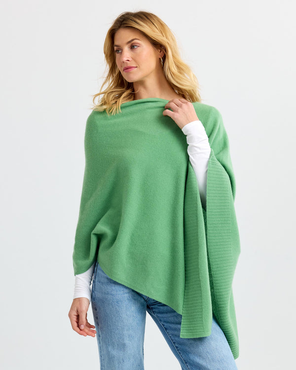 Woman wearing green cashmere poncho over white long sleeve top and blue jeans against plain background