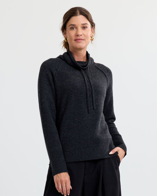 LIFESTYLE / EDITORIAL: Woman wearing charcoal cashmere sweater with cowl neck and drawstrings, standing against white