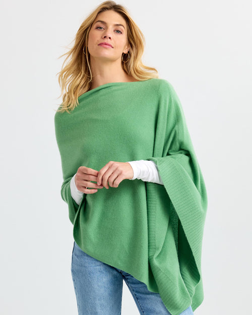 Woman wearing matcha green cashmere poncho over white top and jeans against plain background