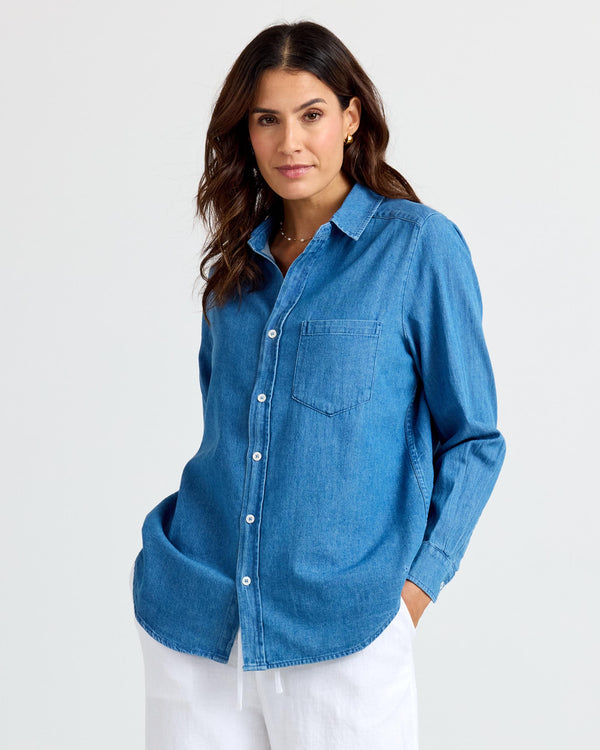 Woman wearing medium wash denim shirt with white pants, standing against plain light background
