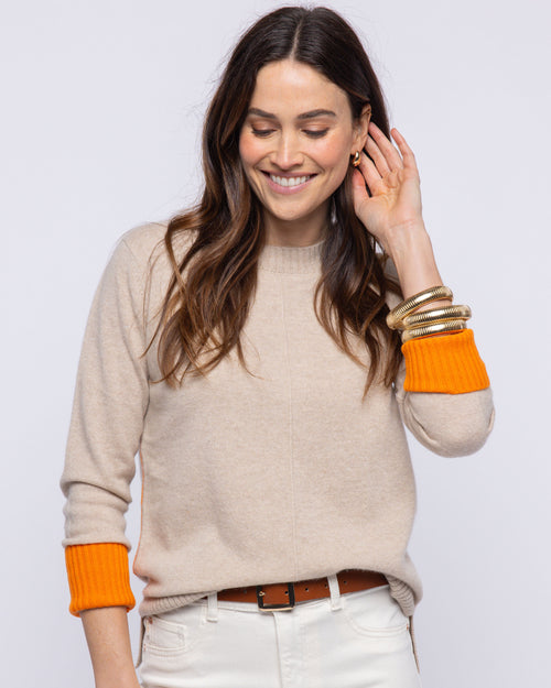 LIFESTYLE / EDITORIAL: Woman wearing beige cashmere crewneck sweater with orange cuffs and white pants, smiling and touching
