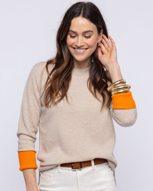 LIFESTYLE / EDITORIAL: Woman wearing beige cashmere crewneck sweater with orange cuffs and white pants, smiling and touching
