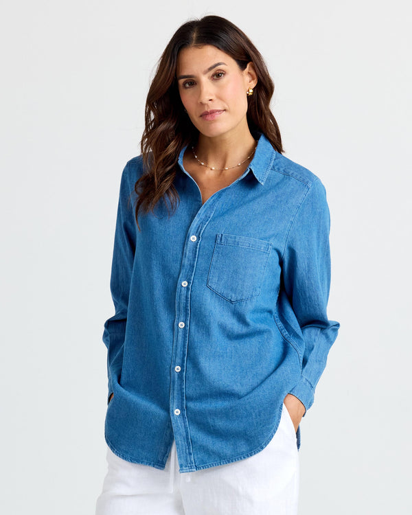 Front-facing image of a woman wearing medium wash denim shirt with white pants, hands in pockets against plain background