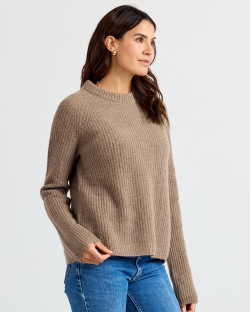 LIFESTYLE / EDITORIAL: Woman wearing driftwood-colored ribbed cashmere sweater and blue jeans, standing against white