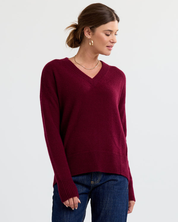 Three-quarter side view image of a woman wearing cabernet red V-neck cashmere sweater and blue jeans against white background