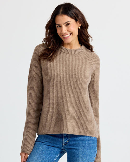 LIFESTYLE / EDITORIAL: Woman smiling wearing a driftwood-colored ribbed cashmere sweater and blue jeans against white