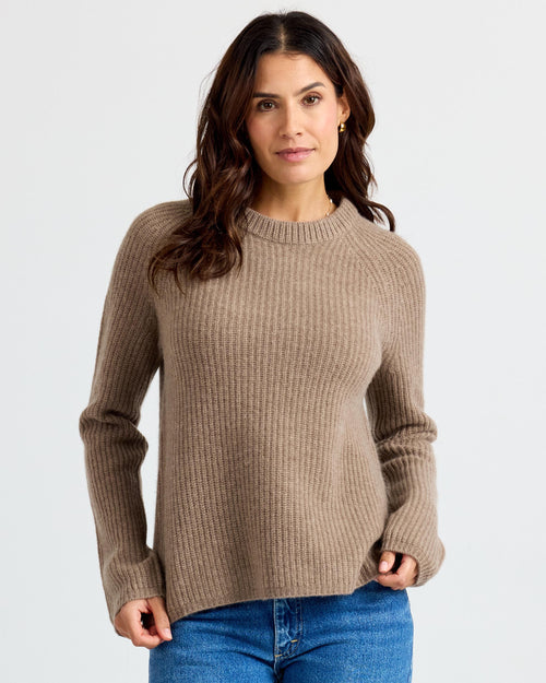 Woman wearing driftwood-colored ribbed cashmere sweater and blue jeans, standing against plain light background