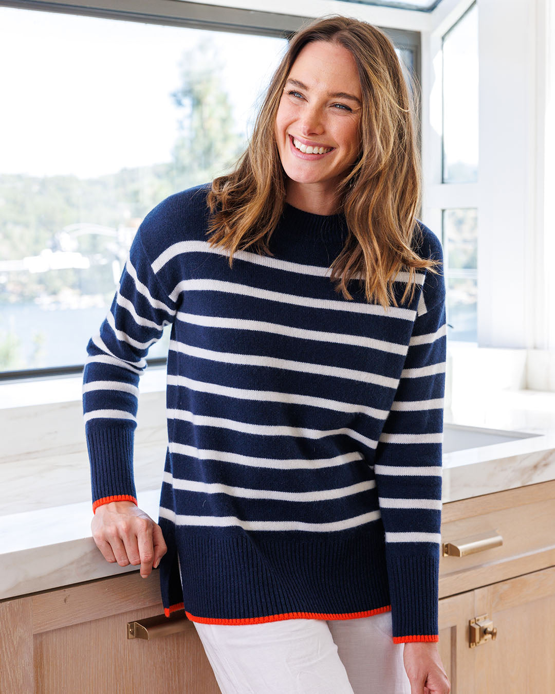 LIFESTYLE / EDITORIAL: Smiling woman in navy and white striped cashmere sweater standing in bright kitchen