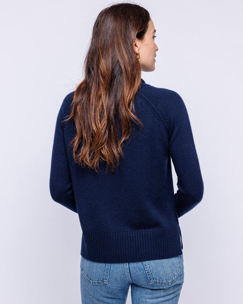Woman wearing navy cashmere sweater and blue jeans, shown from back against plain light background
