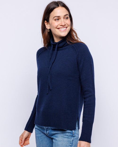 LIFESTYLE / EDITORIAL: Woman wearing navy blue cashmere drawstring funnel neck sweater and jeans, smiling and looking