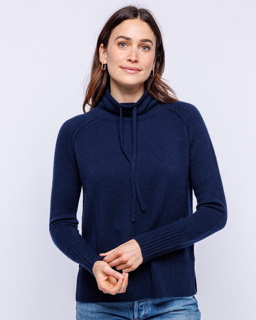 Woman wearing navy blue cashmere drawstring funnel neck sweater with long sleeves and ribbed cuffs, standing against white