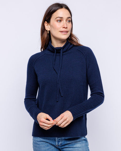 LIFESTYLE / EDITORIAL: Woman wearing navy blue cashmere funnel neck sweater with drawstring, standing against white