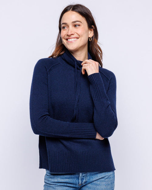 LIFESTYLE / EDITORIAL: Woman wearing navy blue cashmere drawstring funnel neck sweater with jeans, smiling and looking