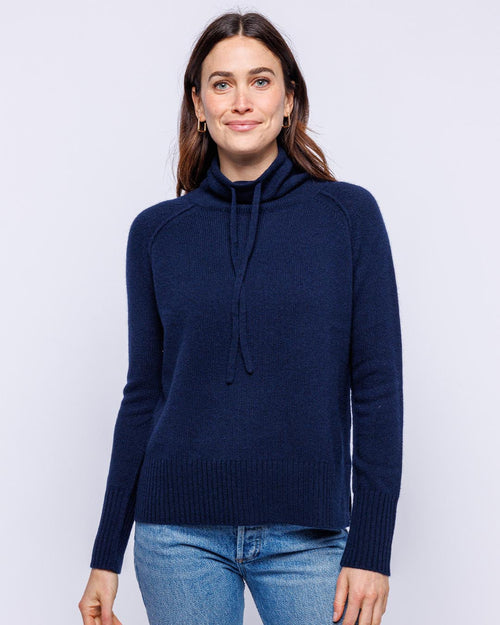 LIFESTYLE / EDITORIAL: Woman wearing navy blue cashmere drawstring funnel neck sweater and jeans against white background