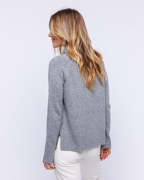 Woman wearing light gray cashmere sweater with drawstring funnel neck, shown from back, paired with white pants