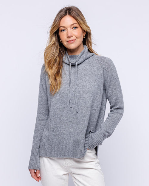 LIFESTYLE / EDITORIAL: Woman wearing grey cashmere drawstring funnel neck sweater with white pants against white background