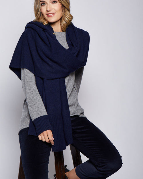 LIFESTYLE / EDITORIAL: Woman seated wearing navy ribbed cashmere wrap over gray sweater and dark pants against plain