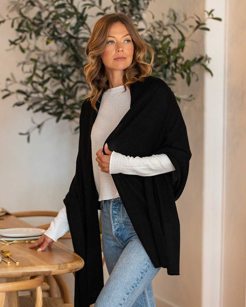 LIFESTYLE / EDITORIAL: Woman wearing black cashmere wrap over white top and jeans, standing by wooden table indoors