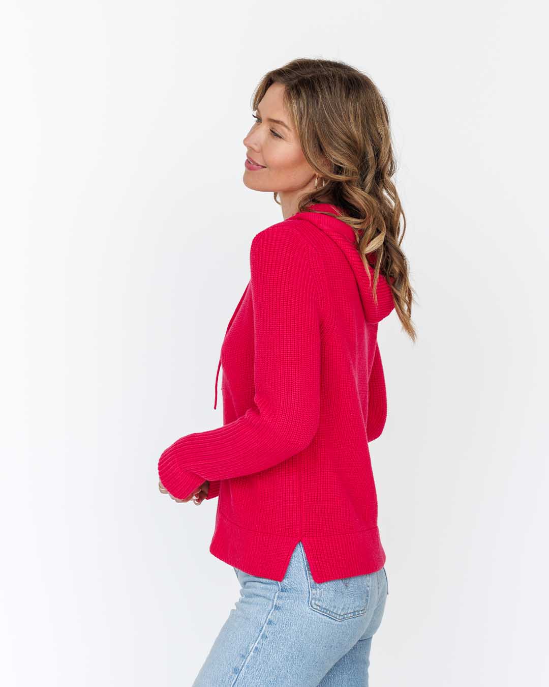 LIFESTYLE / EDITORIAL: Woman wearing bright red hooded sweater and light blue jeans, posing sideways against white background