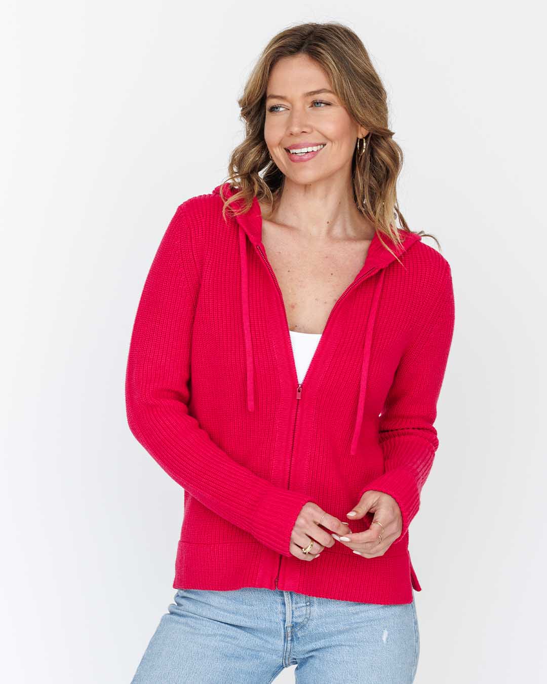 Woman wearing bright pink zip-up cotton hoodie and light blue jeans, smiling, against white background