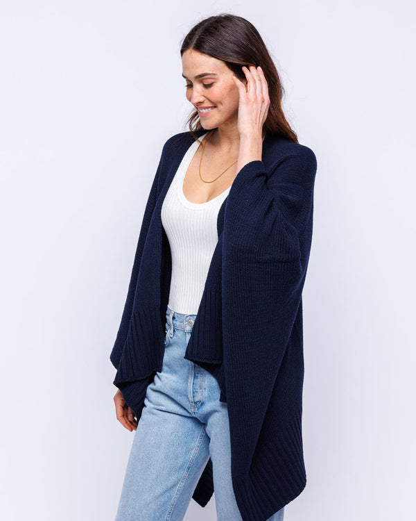 LIFESTYLE / EDITORIAL: Woman wearing navy cotton serape cardigan over white tank and light blue jeans, smiling and touching