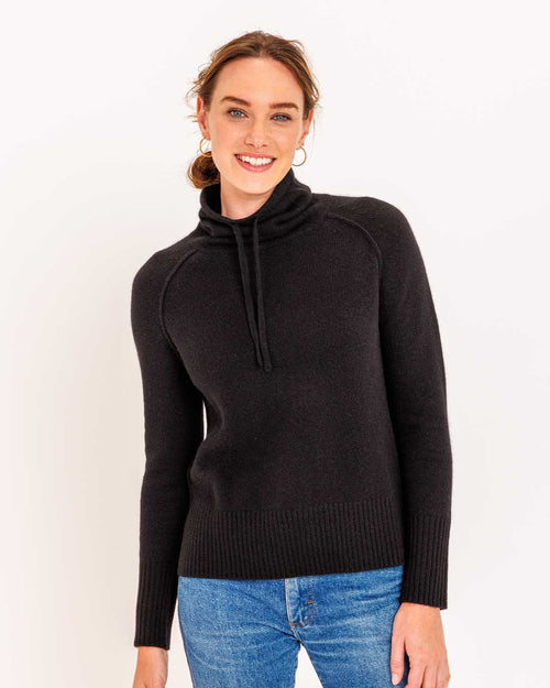 LIFESTYLE / EDITORIAL: Woman wearing black drawstring funnel neck cashmere sweater and blue jeans, smiling against white