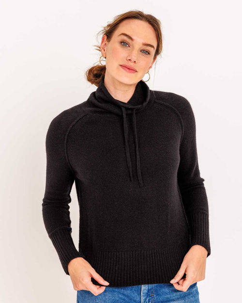 LIFESTYLE / EDITORIAL: Woman wearing black drawstring funnel neck cashmere sweater and blue jeans against white background