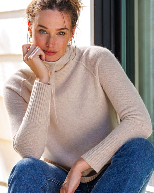 LIFESTYLE / EDITORIAL: Woman seated wearing oatmeal drawstring funnel neck cashmere sweater and blue jeans