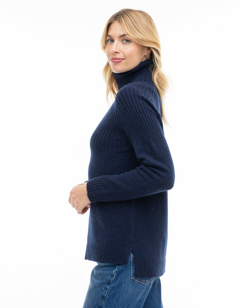 Woman wearing navy blue half-rib turtleneck cashmere sweater, side view, paired with blue jeans against white background