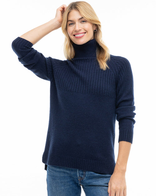 Woman wearing navy half-rib turtleneck cashmere sweater and blue jeans, smiling with hand on head