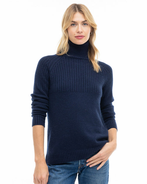 LIFESTYLE / EDITORIAL: Woman wearing navy half-rib turtleneck cashmere sweater and blue jeans, standing against white