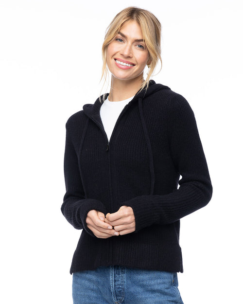 Woman wearing black zip-up cashmere hoodie over white shirt and blue jeans, smiling against white background