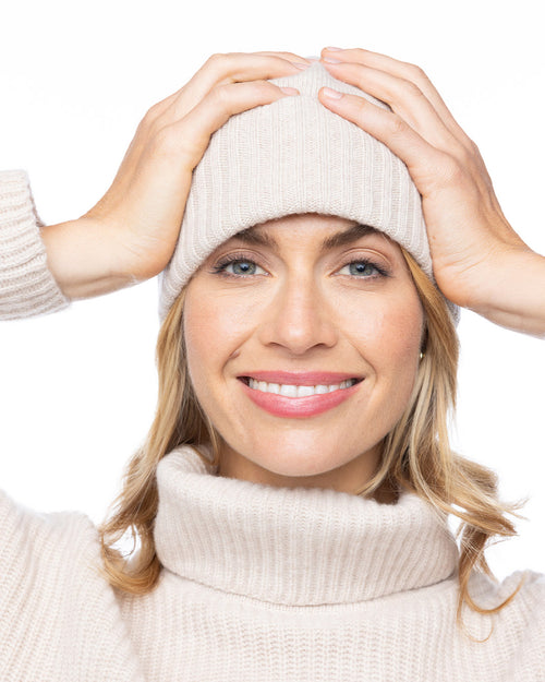 LIFESTYLE / EDITORIAL: Woman smiling in oatmeal-colored cashmere turtleneck and matching beanie, hands on head