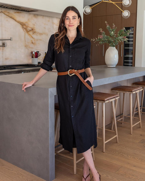 LIFESTYLE / EDITORIAL: Woman in black linen shirt dress with brown belt standing by kitchen island with stools and vase of
