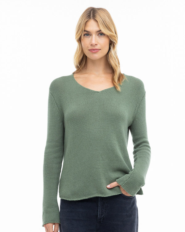Woman wearing olive green round-edge cashmere sweater with long sleeves and slight V-neck, standing against white background