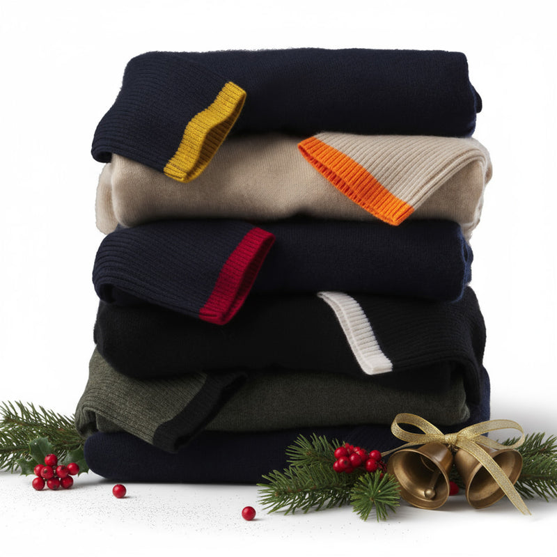 Stack of folded cashmere sweaters with colored cuffs, decorated with pine branches, red berries, and golden bells.