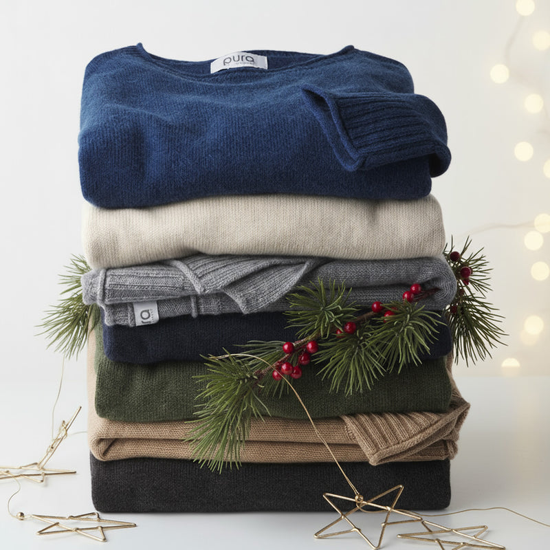 Stack of folded cashmere sweaters in various colors with pine and red berry holiday decorations