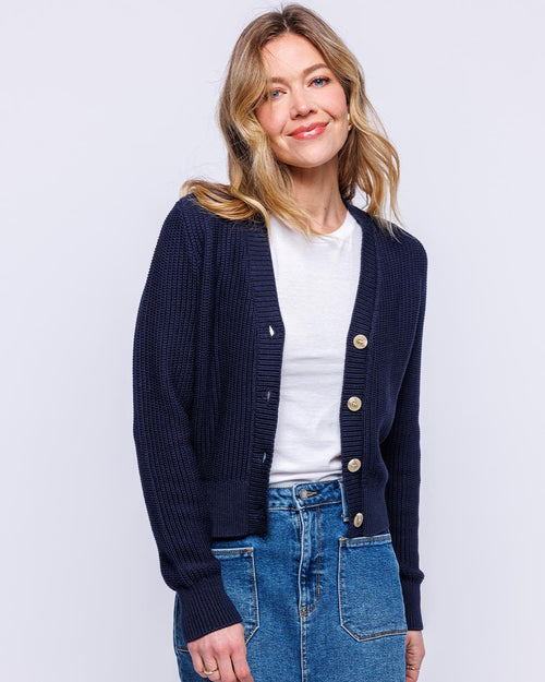 LIFESTYLE / EDITORIAL: Woman wearing navy textured cardigan over white shirt and blue jeans, standing against light